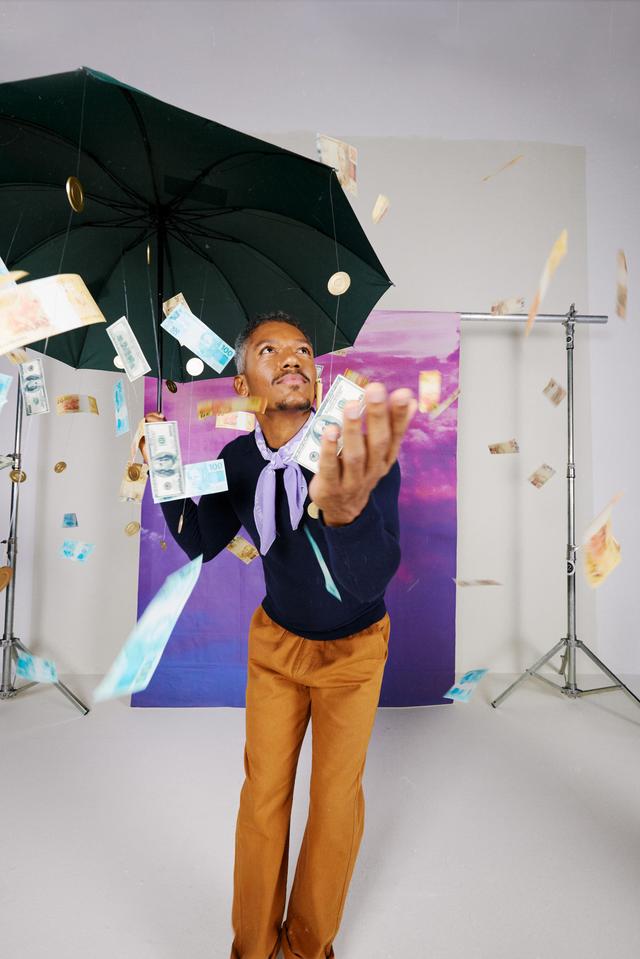 Imagem de um homem negro em um estúdio de fotografia, segurando um guarda-chuva. Moedas e notas de 50 e 100 reais caem sobre ele. Ele está usando uma blusa azul escura, calças marrons e um lenço roxo amarrado ao pescoço. No fundo do estúdio, há um pano pendurado, com um céu cheio de nuvens roxas.