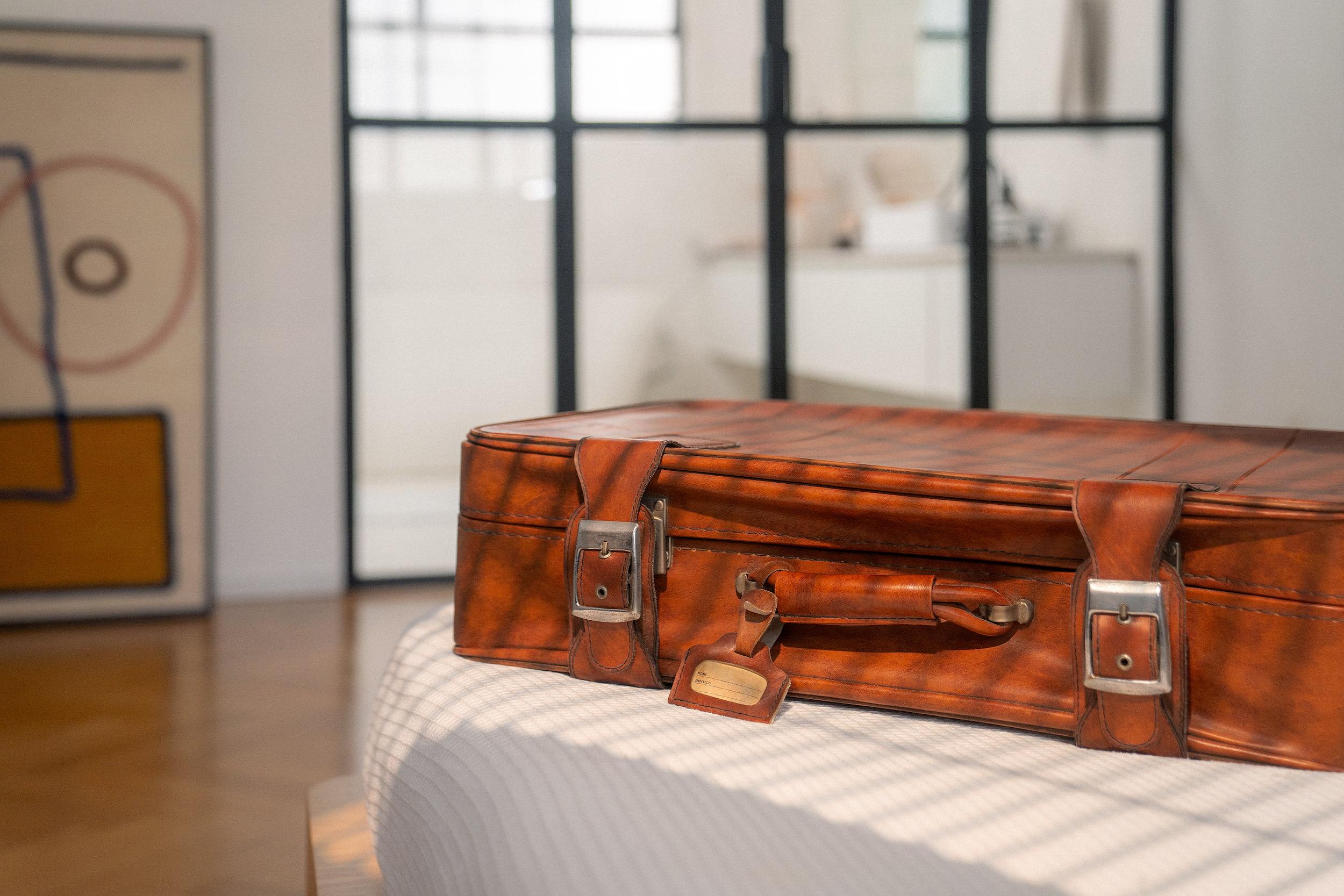 Passagem aérea na Black Friday: imagem de uma mala de viagem sobre uma cama com lençol branco. A mala é marrom, de couro.
