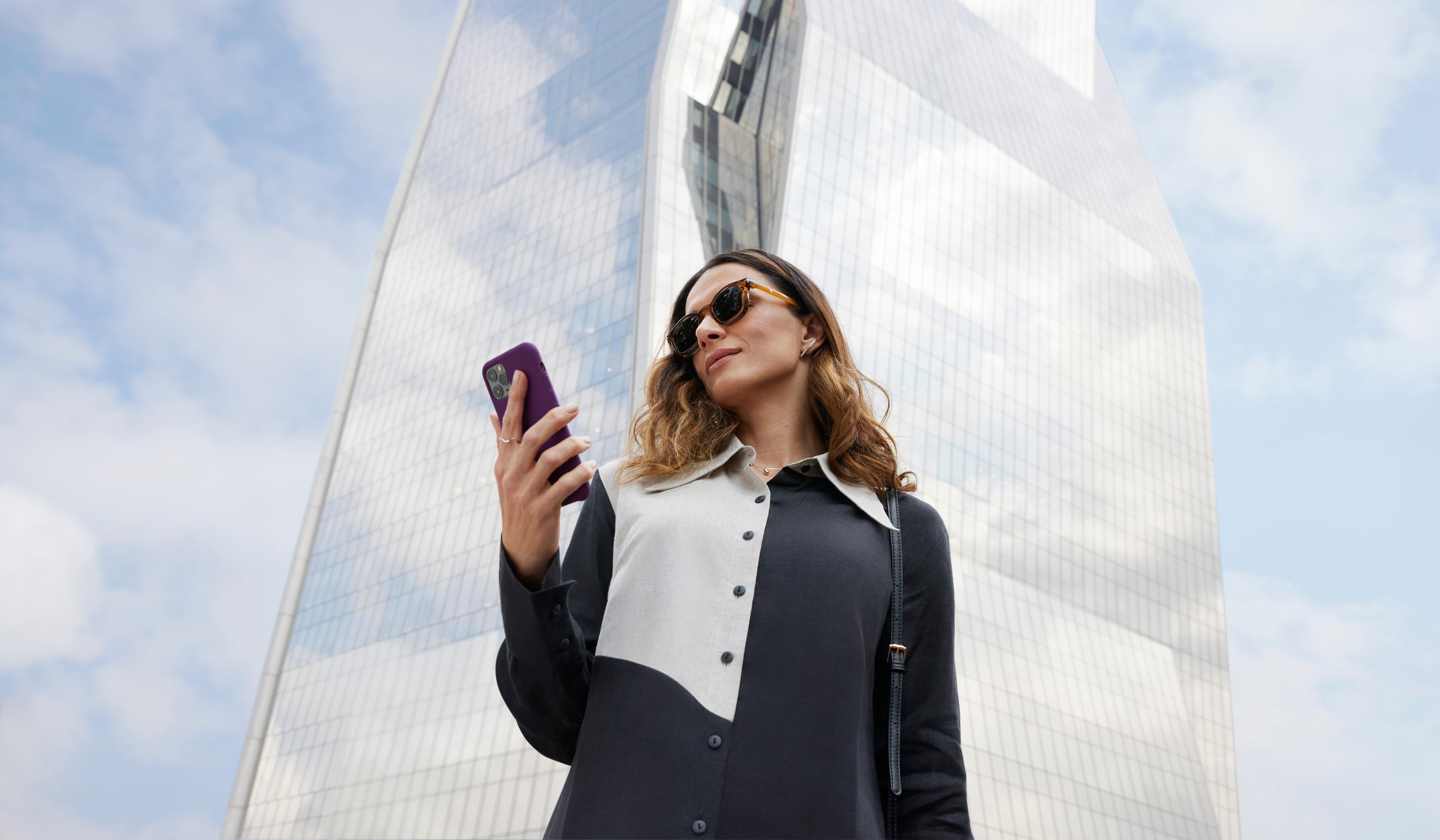 Foto de uma mulher branca, usando óculos de sol. Ela está vestindo uma roupa preta e branca, segurando um celular na mão. Atrás, um prédio espelhado.